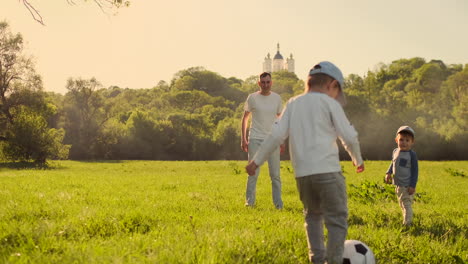 A-young-father-in-a-white-t-shirt-with-two-sons-playing-football-on-the-grass-at-sunset-in-the-sun-in-slow-motion